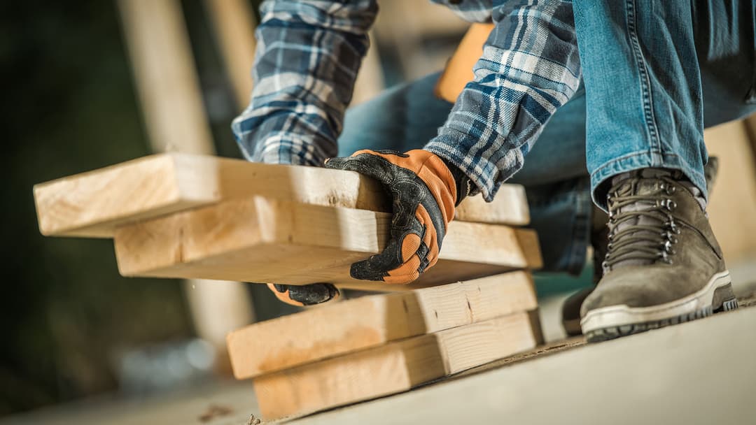 Mies nostaa puutavaraa lattialta, kädessään oranssi-mustat työhanskat