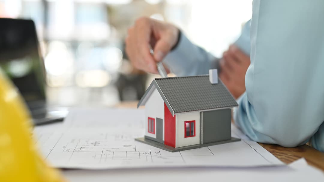 omakotitalon pienoismalli rakennuspiirustusten päällä taustalla sumealla suunnittelijan käsivartta sekä tietokone