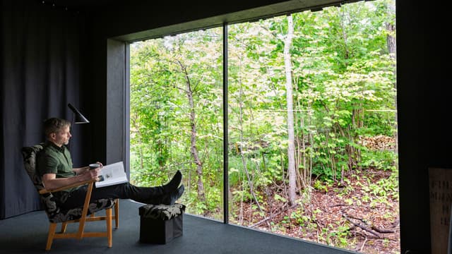 matti kuittinen istuu olohuoneessa jonka koko seinän kokoisesta ikkunasta näkyy vehreä maisema. Huoneessa ei ole kuin nojatuoli ja jalkalamppu